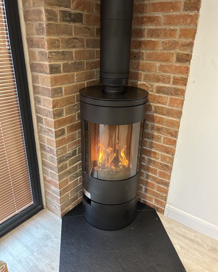 A cylindrical wood burner fitted against an exposed brick chimney breast with the fire lit
