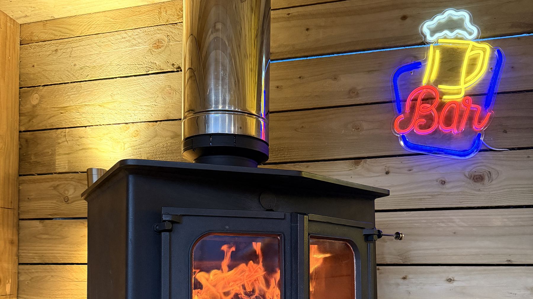 A cast-iron wood burning stove with the fire lit, fitted with a stainless twin-wall flue in a timber-lined garden room