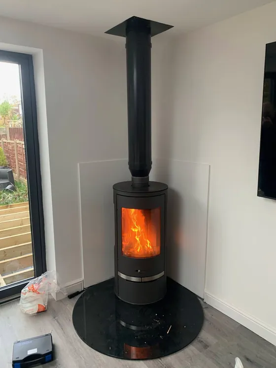 Contemporary cylindrical wood burner on a curved glass hearth, fire lit, beside full-height windows