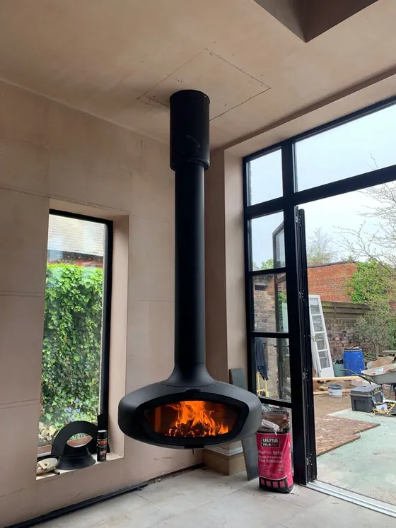 Suspended cylindrical designer stove in an open-plan room with steel-framed glazing, fire burning
