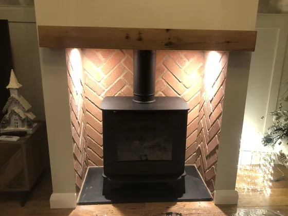 Wood burner in a herringbone brick recess beneath an oak beam with warm uplighting